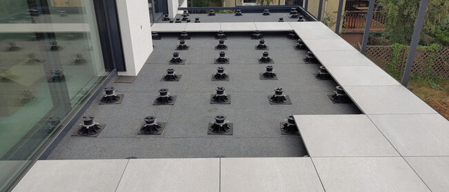 Montage de terrasse surélevée avec dalles en grès cérame gris clair sur plots réglables noirs.