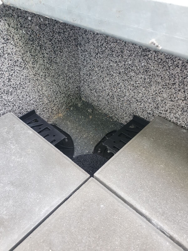 Placement en angle de plots de terrasse découpés supportant des dalles de sol extérieures surélevées contre un mur texturé.