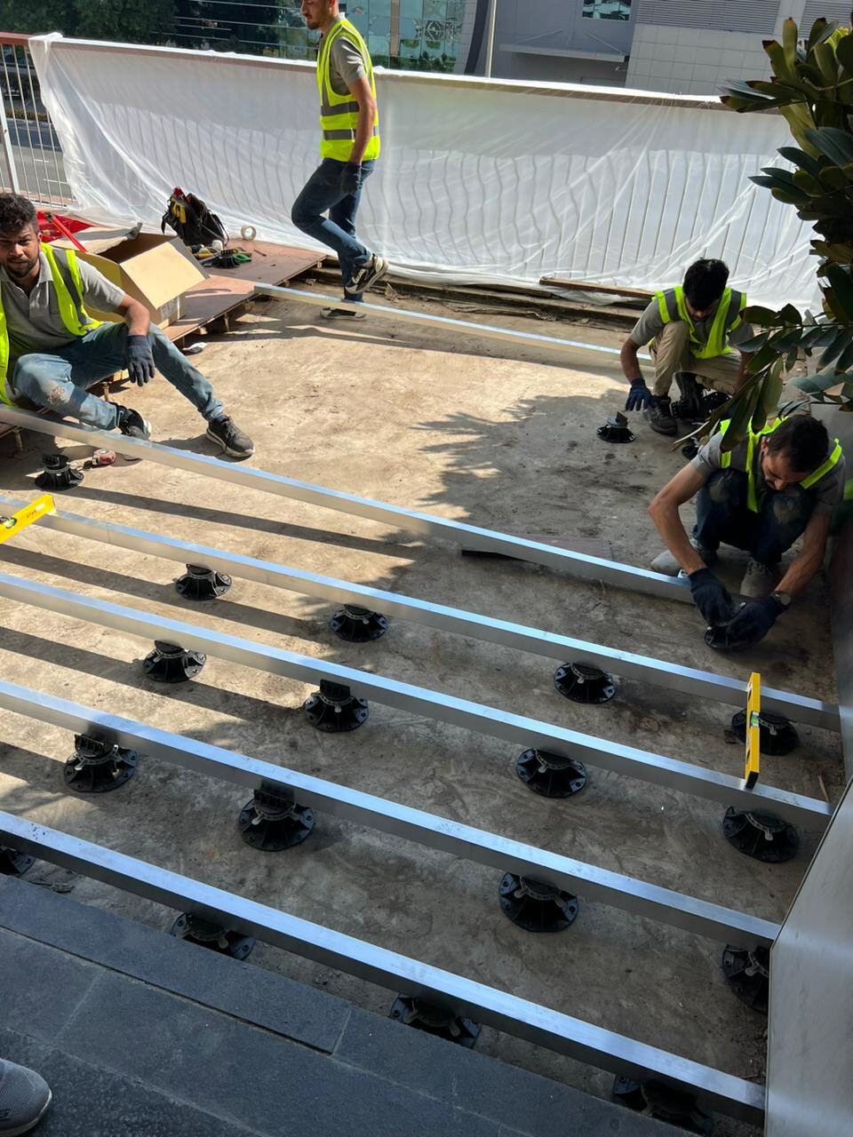 Construction workers installing an outdoor raised decking substructure with aluminum joists and adjustable pedestals.