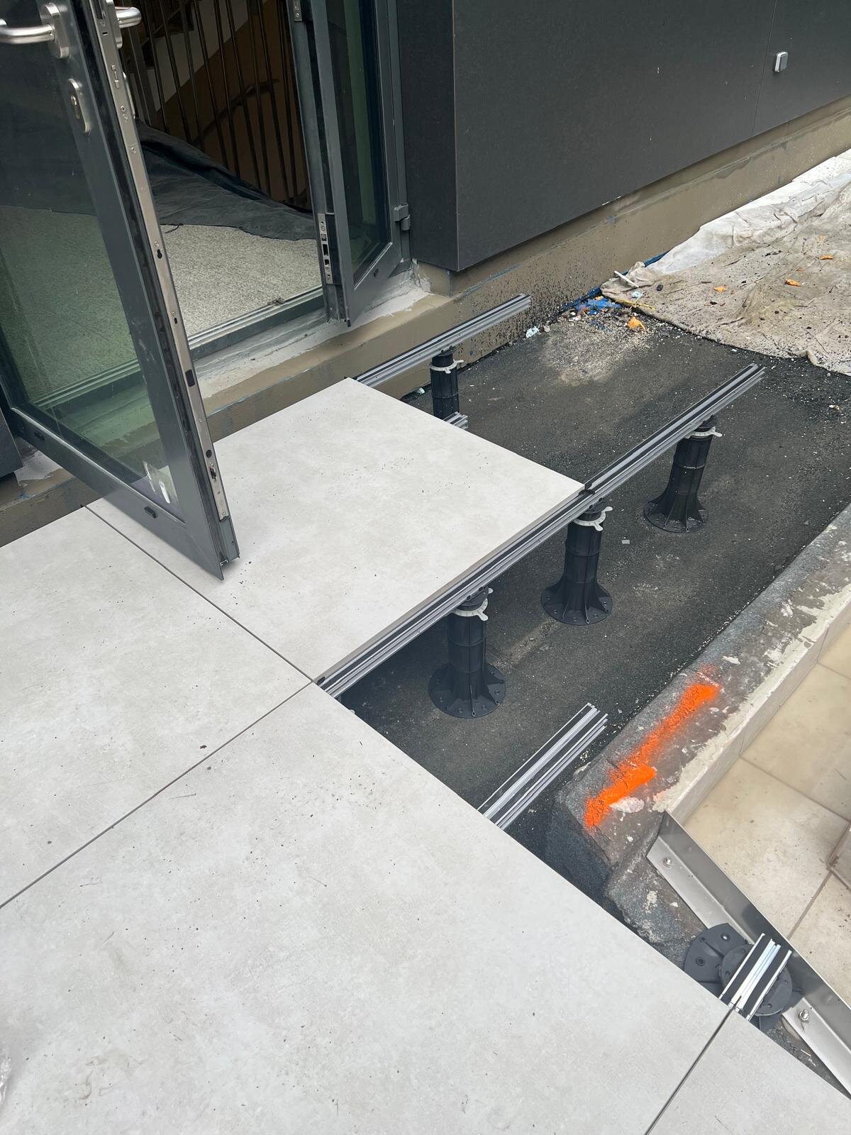 Installation au niveau de la porte de terrasse avec des dalles en porcelaine extérieures posées sur des lambourdes aluminium et des plots réglables en plastique pour terrasse.