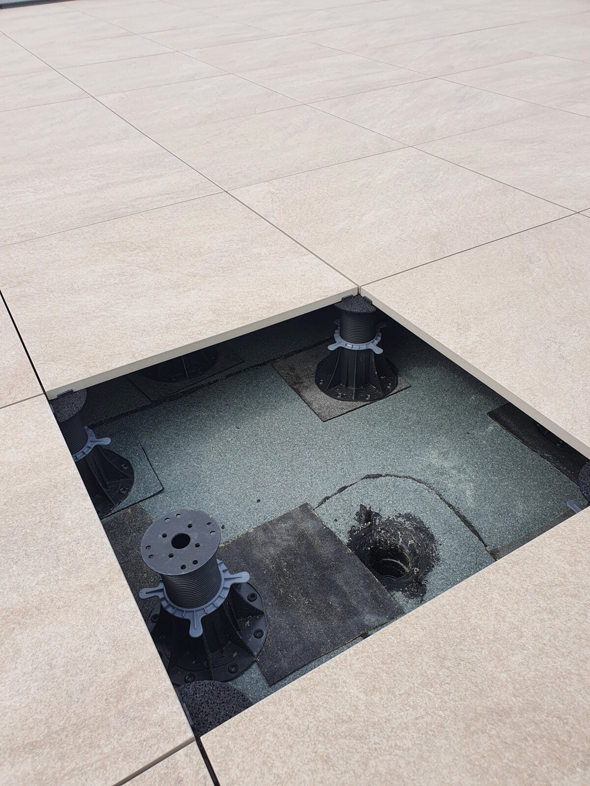 Vue de dessus d'une section de dalle de terrasse ouverte révélant un plot réglable et une évacuation d'eau de toit en dessous.