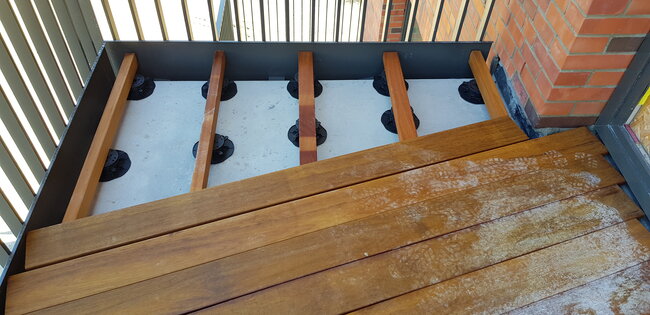 Installation de lames de balcon avec lambourdes en bois visibles soutenues par des plots réglables de terrasse sur une base en béton.