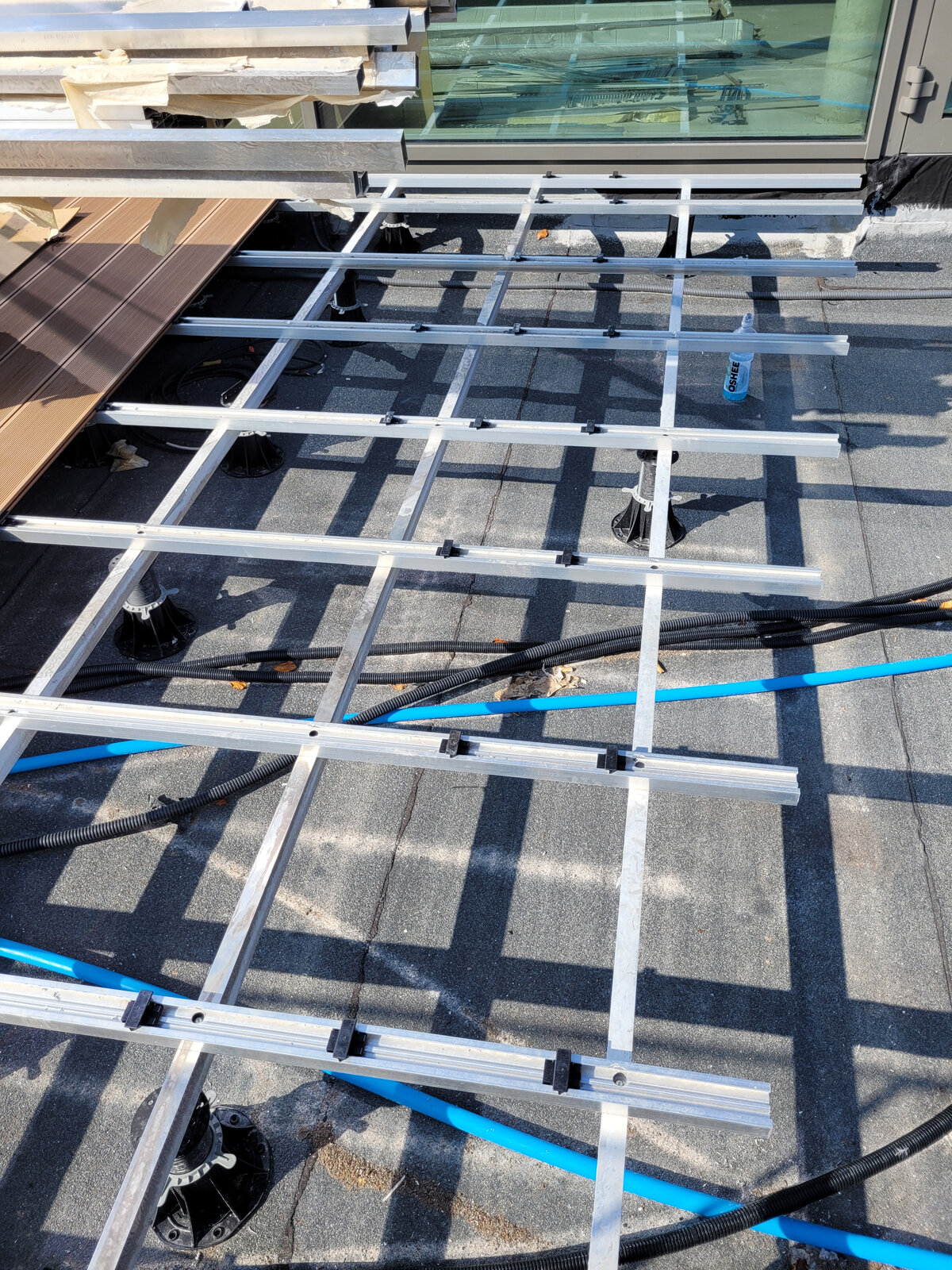 Structure de lambourdes en aluminium montée sur plots réglables, acheminée au-dessus des tuyaux et du câblage sous une terrasse surélevée.