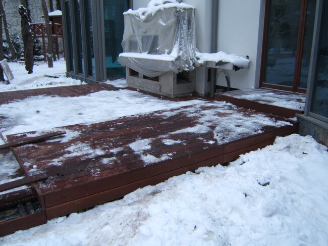 Terrasse en bois foncé partiellement couverte de neige fondante devant une maison en hiver.
