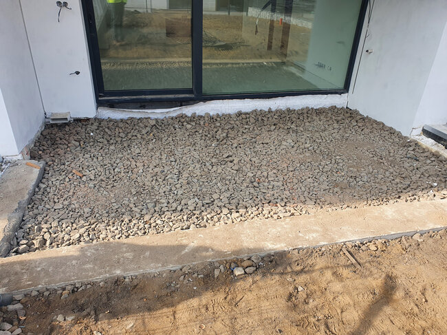Couche de pierre concassée préparée pour la construction d'une terrasse à l'extérieur devant des portes coulissantes en verre.