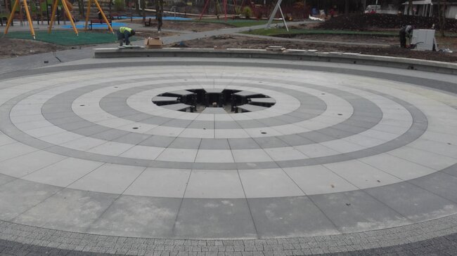 Agencement circulaire de dalles surélevées autour d'une fontaine sèche extérieure soutenu par des plots réglables.